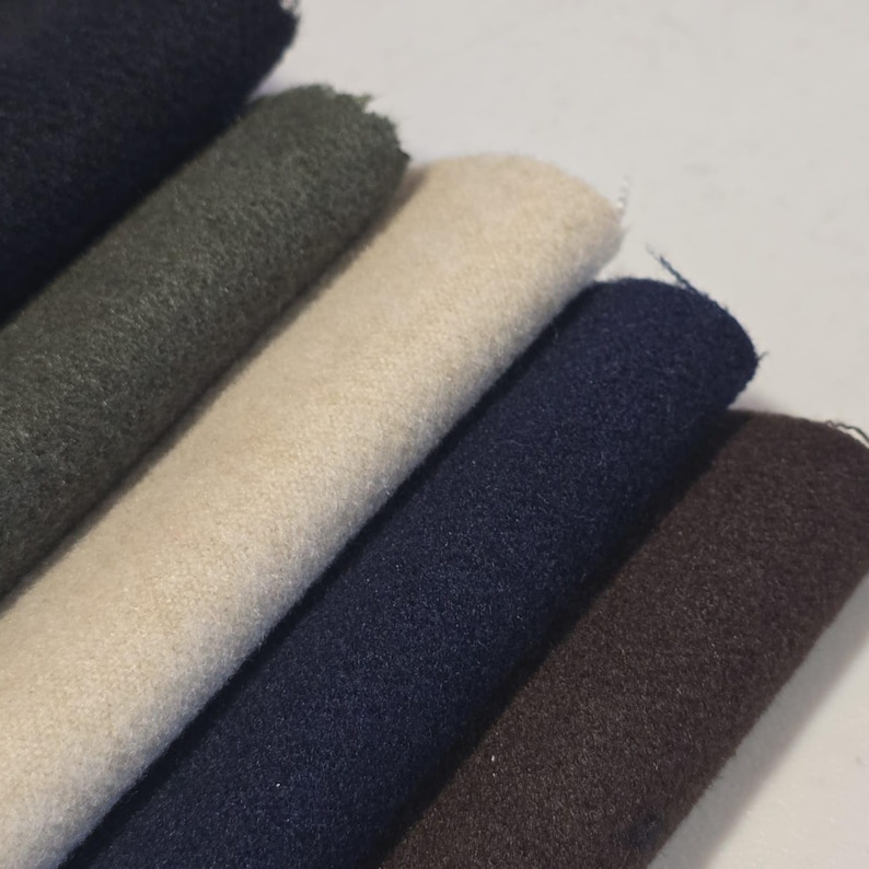 K&ouml;nnte beinhalten: Nahaufnahme von mehreren gerollten Stoffmustern in verschiedenen Farben. Die Farben umfassen Schwarz, Olivgr&uuml;n, Creme, Marineblau und Braun. Der Stoff scheint ein strukturiertes Material zu sein, m&ouml;glicherweise Wolle oder ein &auml;hnliches Textil.