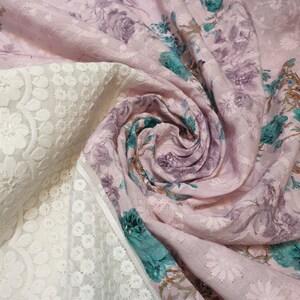Può includere: Tessuto rosa con un motivo floreale e un bordo di pizzo bianco. Il tessuto è piegato e attorcigliato, mostrando la trama e il dettaglio del design.