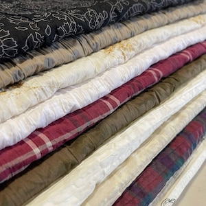 May include: A selection of folded fabric textiles in various colours and patterns. The fabrics include a black floral print, beige, white, and burgundy plaid. Some fabrics have a crinkled texture, others are smooth.