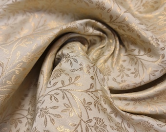 Op de afbeelding: Een close-up van een crèmekleurige stof met een gouden bloemenpatroon. De stof is zacht en luxueus, en het patroon is ingewikkeld en elegant.