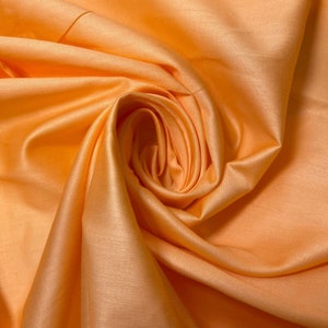 May include: A close-up of a peach-colored fabric with a smooth, silky texture. The fabric is folded and draped, creating soft, flowing curves.