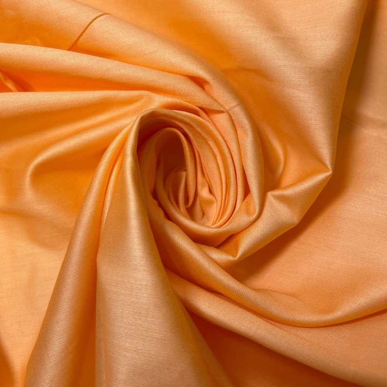 May include: A close-up of a peach-colored fabric with a smooth, silky texture. The fabric is folded and draped, creating soft, flowing curves.