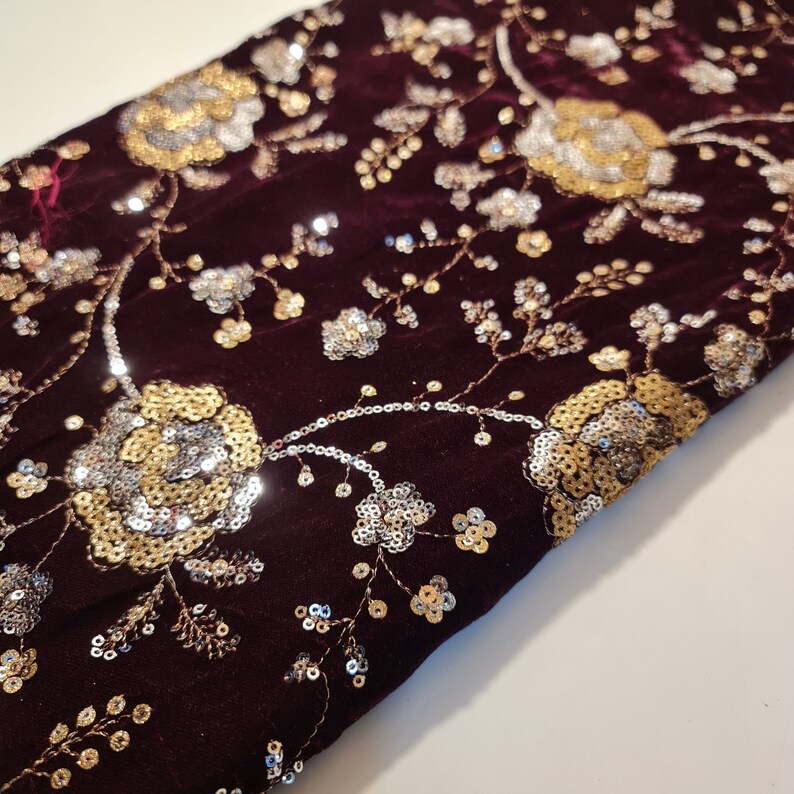 K&ouml;nnte beinhalten: Burgunderfarbener Samtstoff mit goldenen und silbernen Pailletten in einem Blumenmuster.