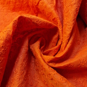 Könnte beinhalten: Orangener Stoff mit einem strukturierten Blumenmuster. Der Stoff ist gefaltet und drapiert und erzeugt einen weichen und fließenden Look.
