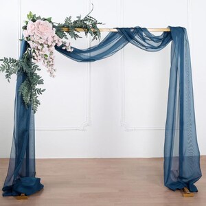 May include: A gold metal archway decorated with a teal blue sheer fabric and a cascading garland of pink flowers and greenery.