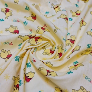 May include: Yellow fabric with a repeating pattern of cartoon Winnie the Pooh bears. The bears are wearing red shirts and are in various poses. The background is a light yellow colour.