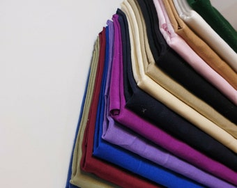 Op de afbeelding: Een stapel stofstalen in verschillende kleuren, waaronder groen, blauw, paars, rood, beige en zwart. De stof is netjes gevouwen en in een driehoekige vorm gerangschikt.