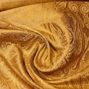 Op de afbeelding: Een gouden stof met een paisley-patroon. De stof is zacht en heeft een subtiele glans.