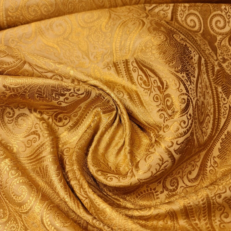 Op de afbeelding: Een gouden stof met een paisley-patroon. De stof is zacht en heeft een subtiele glans.