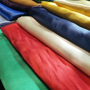 Può includere: Un rotolo di tessuto di raso colorato in varie tonalità di verde, rosso, bianco, beige, blu, giallo e marrone. Il tessuto è liscio e lucido, e i rotoli sono impilati ordinatamente uno sopra l'altro.
