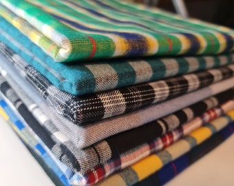 Puede incluir: Una pila de muestras de tela a cuadros en varios colores, incluyendo verde, azul, negro y amarillo. La tela está doblada cuidadosamente y dispuesta en forma de pirámide.