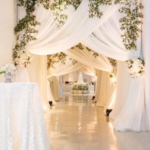 May include: A white wedding ceremony aisle decorated with white fabric draping and greenery. The aisle leads to a white wedding cake decorated with white flowers.