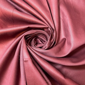 May include: A close-up of a deep burgundy fabric with a smooth, silky texture. The fabric is folded and draped in a way that creates a swirling, rose-like pattern.