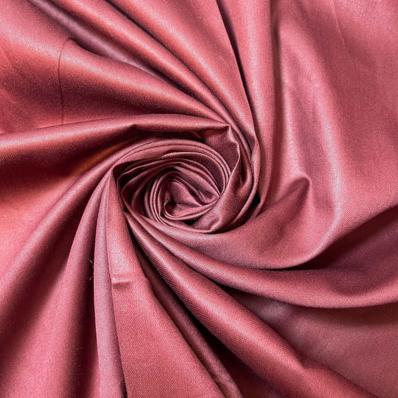 May include: A close-up of a deep burgundy fabric with a smooth, silky texture. The fabric is folded and draped in a way that creates a swirling, rose-like pattern.