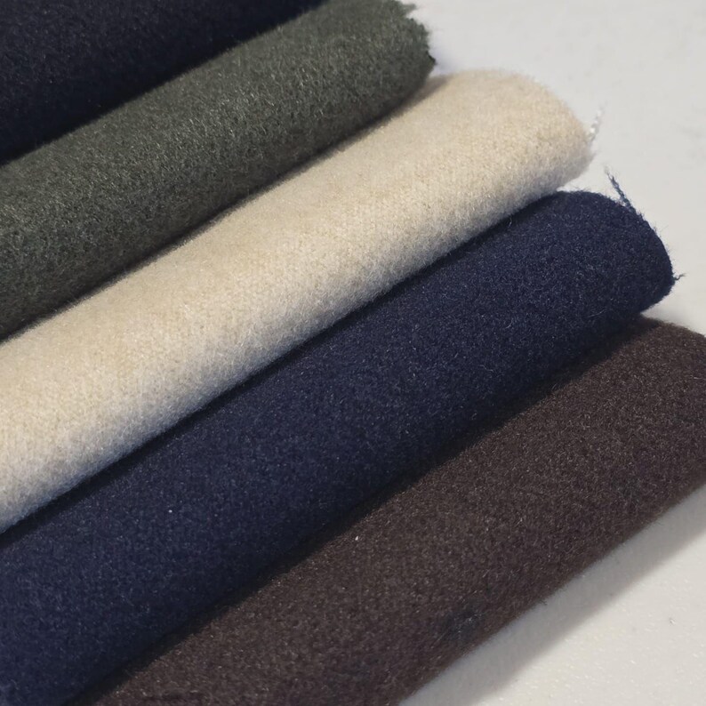 K&ouml;nnte beinhalten: Nahaufnahme von mehreren Stoffrollen in verschiedenen Farben. Die Farben umfassen Dunkelmarine, Olivgr&uuml;n, Creme, Marineblau und Braun. Der Stoff scheint ein weiches, strukturiertes Material zu sein, m&ouml;glicherweise Wolle oder eine Wollmischung.