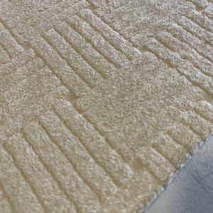 May include: Close-up of a textured, beige fabric with a woven, geometric pattern. The design features a series of parallel lines arranged in a diagonal, creating a textured surface. The fabric appears soft and plush, suitable for upholstery or home decor.