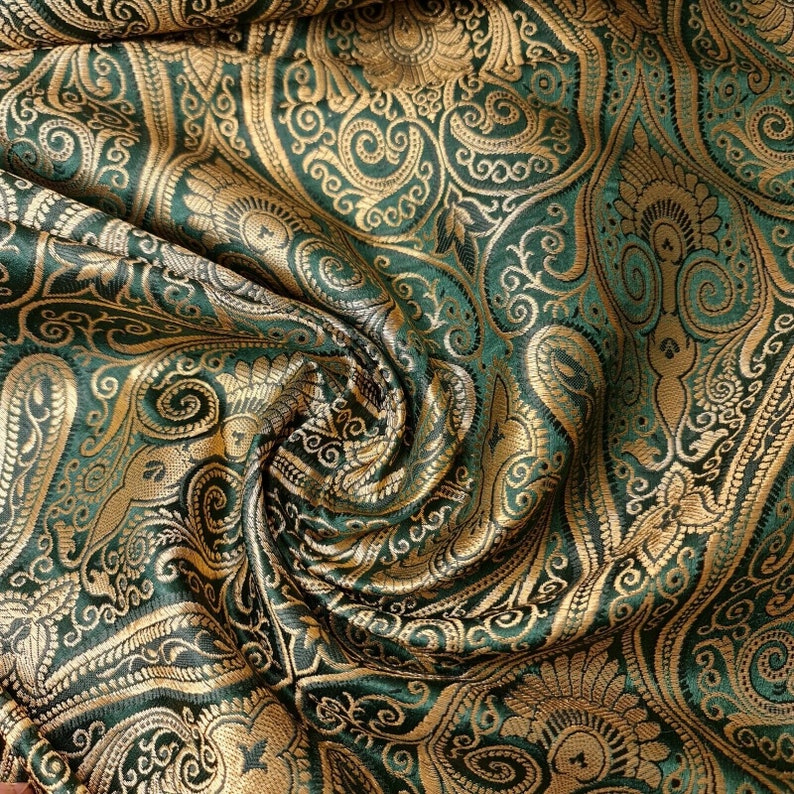 Op de afbeelding: Groene en gouden brokaat stof met een paisley patroon. De stof is gevouwen en gedrapeerd, waardoor de textuur en de details van het ontwerp zichtbaar worden.