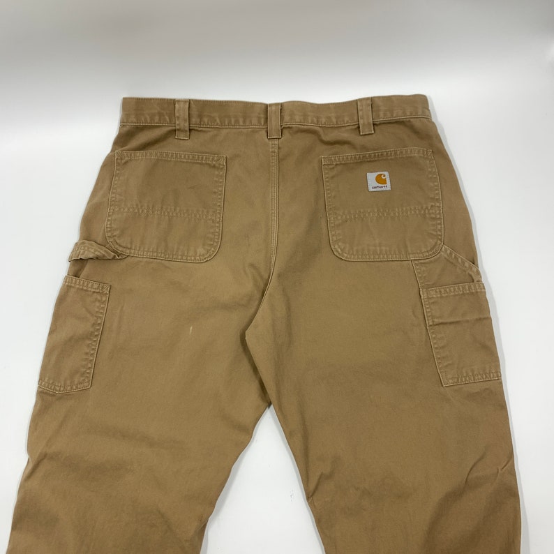 Puede incluir: Pantalones de trabajo color caqui con m&uacute;ltiples bolsillos, incluyendo un bolsillo lateral y dos traseros. El logo de Carhartt es visible en el bolsillo trasero. Los pantalones est&aacute;n hechos de un material duradero.