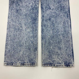 May include: Close-up of acid wash denim jeans. The jeans are a medium blue color with a white, speckled pattern throughout. The bottom hems of the jeans are frayed.