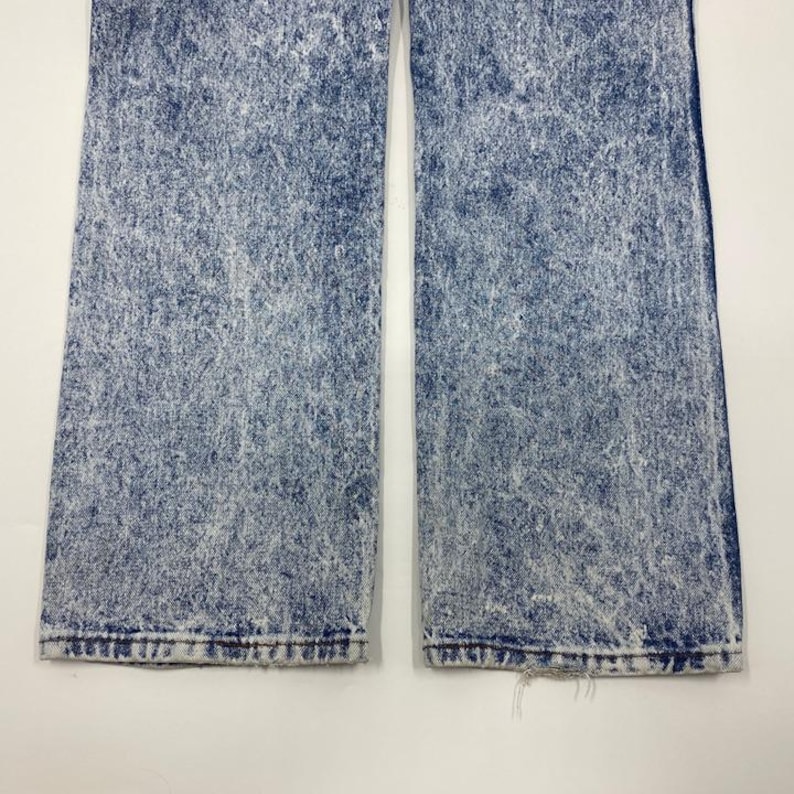 May include: Close-up of acid wash denim jeans. The jeans are a medium blue color with a white, speckled pattern throughout. The bottom hems of the jeans are frayed.