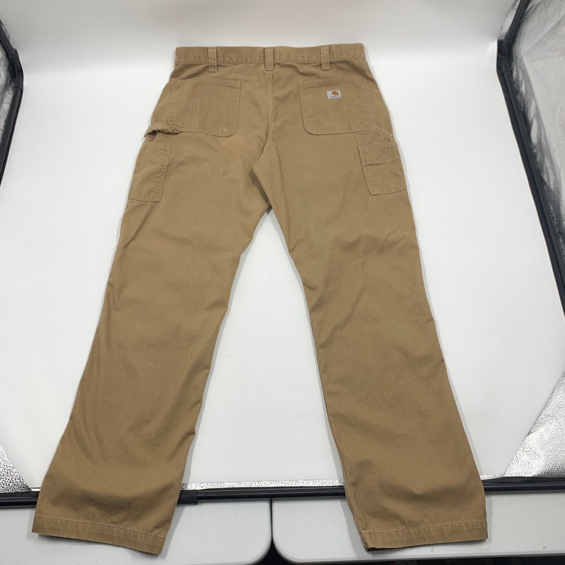Puede incluir: Pantalones de trabajo de color caqui con m&uacute;ltiples bolsillos, incluyendo dos bolsillos traseros y dos bolsillos laterales. Los pantalones tienen un dise&ntilde;o de pierna recta y una etiqueta de la marca Carhartt en el bolsillo trasero. Los pantalones est&aacute;n planos sobre una superficie blanca.