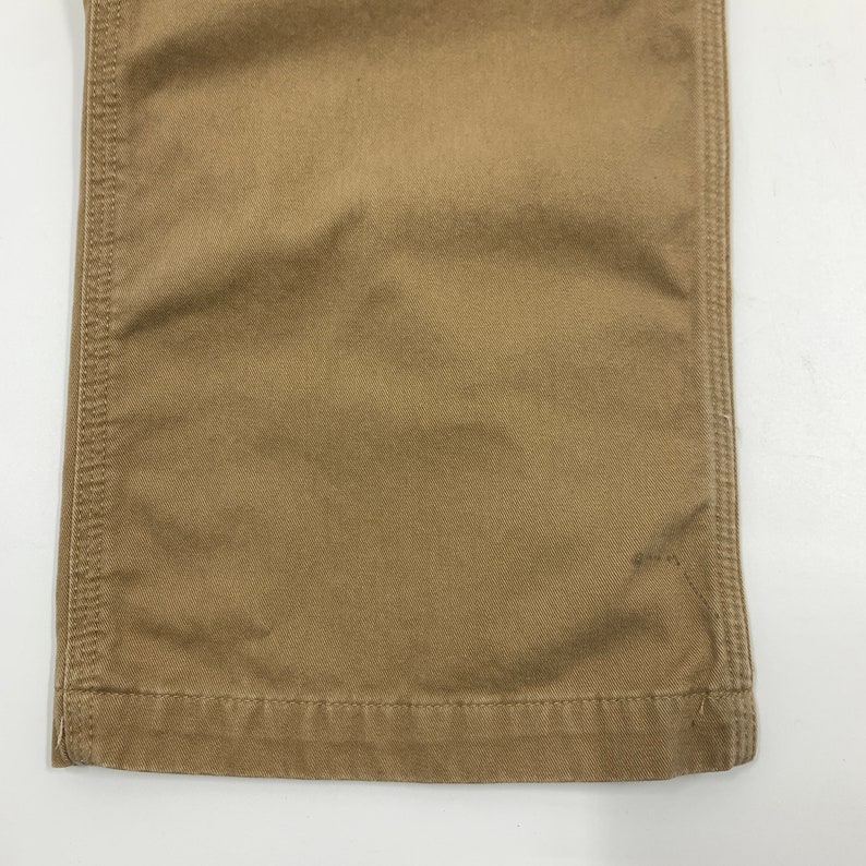 Puede incluir: Primer plano de la parte inferior de un pantal&oacute;n color caqui. La tela es de color beige liso con costuras visibles a lo largo de las costuras y el dobladillo. El pantal&oacute;n parece estar hecho de un material duradero.