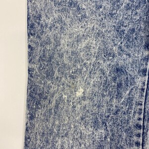 May include: Close-up of acid wash denim fabric. The blue denim has a faded, textured appearance with white highlights and a few light-colored spots. The fabric is folded, showing a seam on the right side.