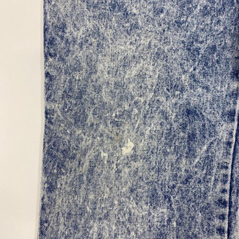 May include: Close-up of acid wash denim fabric. The blue denim has a faded, textured appearance with white highlights and a few light-colored spots. The fabric is folded, showing a seam on the right side.