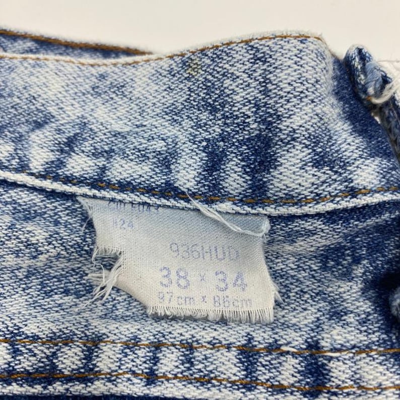 May include: Close-up of a light blue denim jacket with a faded wash. A white tag is attached, displaying the size 38 x 34, and measurements of 97cm x 86cm. The jacket's stitching is visible, and the fabric shows signs of wear.