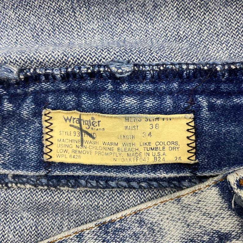 May include: Close-up of a pair of blue denim jeans with a Wrangler brand tag. The tag indicates a waist size of 38 inches and a length of 34 inches. The jeans are made in the USA.