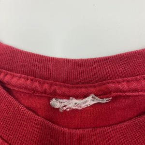Puede incluir: Primer plano de un cuello de camiseta rojo. El cuello es acanalado y muestra signos de desgaste. Una etiqueta blanca rasgada est&aacute; adherida al cuello. La tela parece ser de algod&oacute;n.