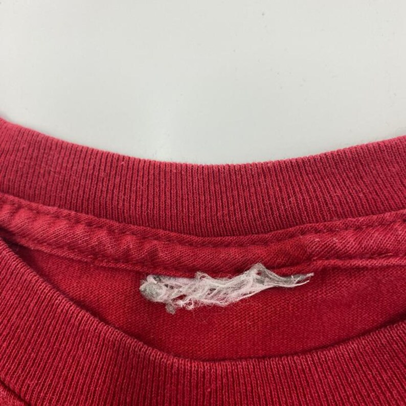 Puede incluir: Primer plano de un cuello de camiseta rojo. El cuello es acanalado y muestra signos de desgaste. Una etiqueta blanca rasgada est&aacute; adherida al cuello. La tela parece ser de algod&oacute;n.