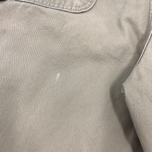 Puede incluir: Primer plano de una tela de color beige con una superficie texturizada. La tela parece ser parte de una prenda, con una costura y un detalle de cuello visibles. Hay una peque&ntilde;a marca blanca en la tela.