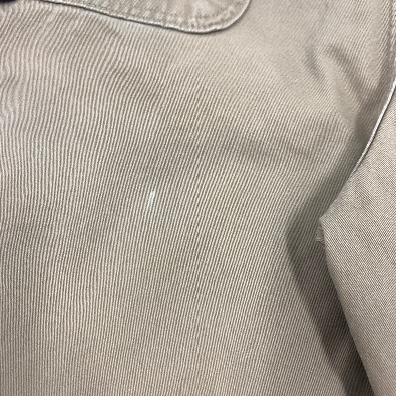 Puede incluir: Primer plano de una tela de color beige con una superficie texturizada. La tela parece ser parte de una prenda, con una costura y un detalle de cuello visibles. Hay una peque&ntilde;a marca blanca en la tela.