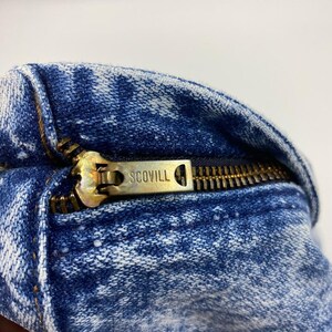 May include: Close-up of a blue denim fabric with a brass zipper. The zipper pull is marked with the brand name "SCOVILL". The denim has a faded, distressed appearance.