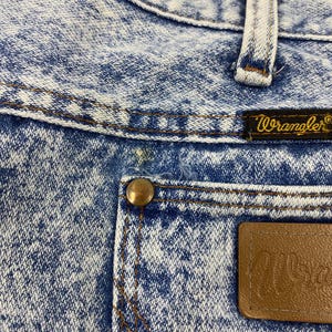 May include: Close-up of acid wash denim jeans with a brown leather patch and a brass button. The jeans feature the brand name "Wrangler" on a small label. The denim has a blue and white pattern.