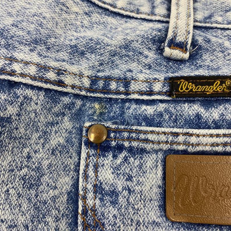 May include: Close-up of acid wash denim jeans with a brown leather patch and a brass button. The jeans feature the brand name "Wrangler" on a small label. The denim has a blue and white pattern.