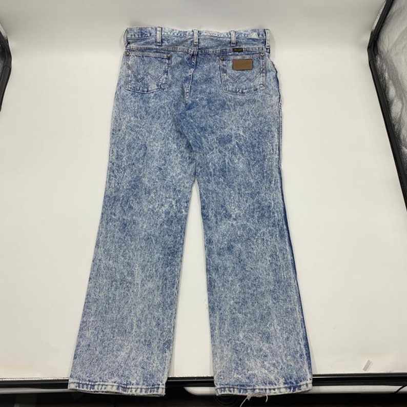 May include: Light blue acid wash denim jeans with a classic five-pocket design. The jeans feature a brown leather patch on the back and a flared leg opening. The denim has a faded, textured appearance.
