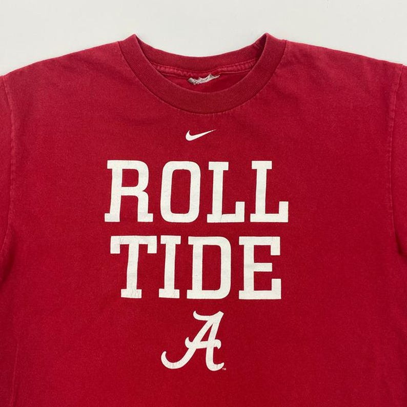 Puede incluir: Camiseta roja con las palabras "ROLL TIDE" en letras blancas grandes y la letra "A" en escritura blanca. Un peque&ntilde;o logotipo blanco de Nike est&aacute; encima del texto. La camiseta tiene cuello redondo.