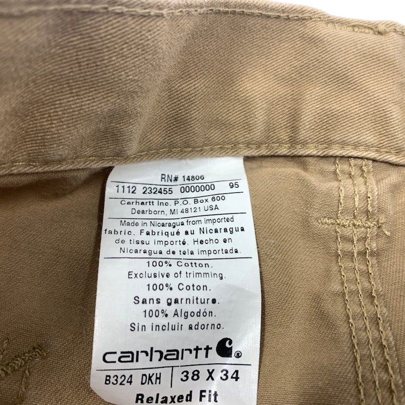 Puede incluir: Primer plano de una etiqueta Carhartt color beige cosida en la cintura de unos pantalones beige. La etiqueta incluye el n&uacute;mero RN, la talla 38 x 34 y el texto "Relaxed Fit". La etiqueta tambi&eacute;n indica que los pantalones est&aacute;n fabricados en Nicaragua.