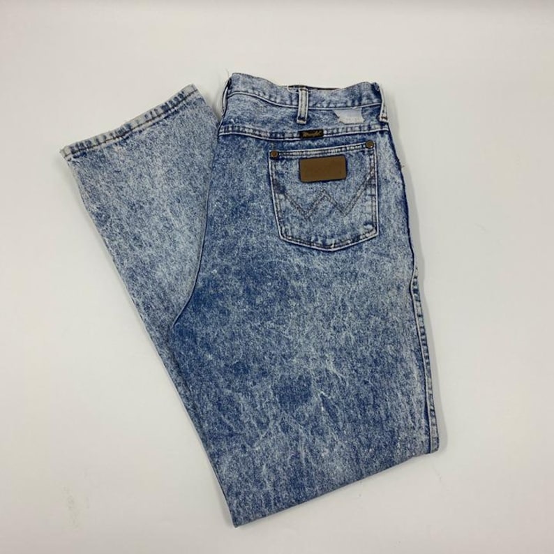 May include: Acid wash denim jeans with a classic five-pocket design. The jeans feature a light blue and white marbled pattern, a brown leather patch, and the Wrangler logo on the back pocket.