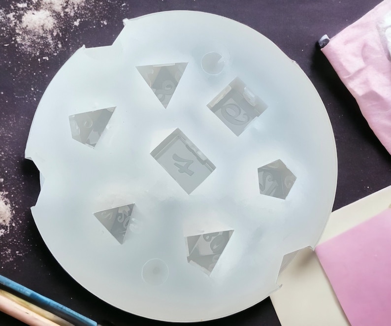 Puede incluir: Un molde de silicona transparente con seis formas diferentes para hacer dados de resina. El molde es circular y tiene una superficie lisa y blanca.