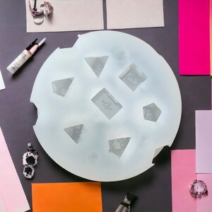 Puede incluir: Un molde de silicona blanco con siete formas diferentes, incluyendo tri&aacute;ngulos, cuadrados y pent&aacute;gonos. El molde est&aacute; sobre una superficie gris con varios cuadrados de papel de colores y otros materiales de artesan&iacute;a.