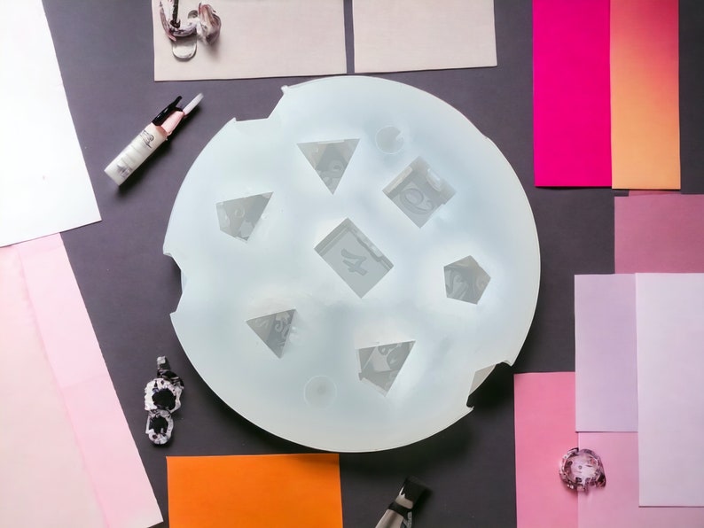 Puede incluir: Un molde de silicona blanco con siete formas diferentes, incluyendo tri&aacute;ngulos, cuadrados y pent&aacute;gonos. El molde est&aacute; sobre una superficie gris con varios cuadrados de papel de colores y otros materiales de artesan&iacute;a.