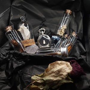 May include: A selection of curiosities in a black box. The items include glass bottles with corks, a small glass dome, a silver pocket watch, and dried plants. The background is black tissue paper.