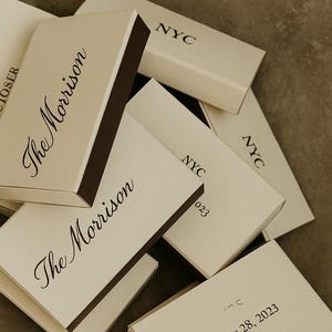 May include: Cream-coloured matchboxes with dark brown edges and text, including "The Morrison," "NYC," and "October 28, 2023." The image also features the text "modern & personalised matchboxes."