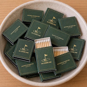 May include: A selection of dark green matchboxes with gold lettering and a golf-themed design. The text "30 UNDER PAR" and "MATTEO'S 30 UNDER PAR" are visible on the boxes. Some boxes are open, revealing the matchsticks. The matchboxes are in a beige bowl.