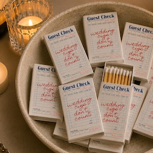 May include: A collection of matchboxes with the text "Guest Check" and "wedding cigars don't count" in a beige bowl. The matchboxes are white with red and blue text. Lit candles and a champagne bottle are in the background.