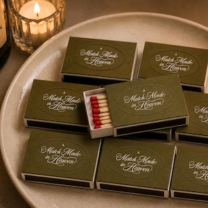 Puede incluir: Cajas de cerillas verde oliva con el texto "Match Made in Heaven" en escritura blanca, dispuestas sobre un plato crema. Una caja está abierta, revelando cerillas con puntas rojas. Velas y una botella de champán en el fondo.