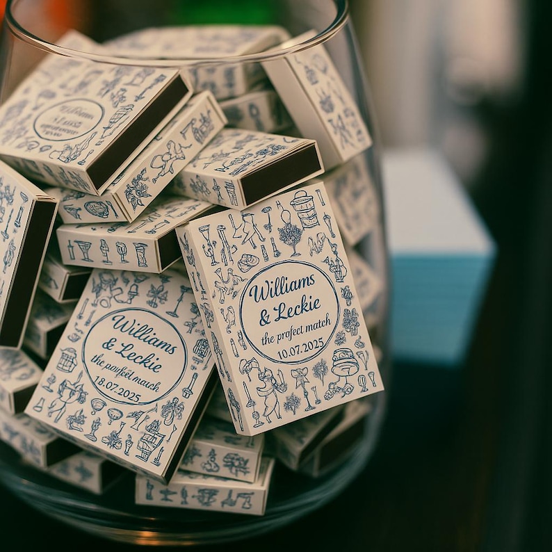 May include: A glass bowl filled with matchboxes. The matchboxes are white with blue illustrations and text that reads "Williams & Leckie the perfect match" with a date. The match heads are dark brown.